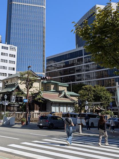 In the middle of the busy city of Kobe, looking straight at a old japanese shrine building across the street intersection, which has wide a wide crosswalk. A few trees grow on shrine grounds and at the side of the street.  People are walking over it and the connected sidewalks. On the street, three cars are moving to the right of the frame. In the background on the left, there's a high rise building and on the right, there two smaller buildings with 8 and about 15 floors respectively.