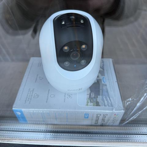 Op de foto zie je een witte beveiligingscamera van het merk EZVIZ. De camera staat binnen achter een raam en is gericht naar buiten. Hij staat op de originele doos van het product. De camera heeft een ronde vorm en een zwart voorpaneel met vier kleine lampjes rond de lens. Er branden drie blauwe lampjes op de camera, wat waarschijnlijk aangeeft dat hij actief is. Op de doos onder de camera staat “Smart Home Camera” en zijn verschillende functies afgebeeld, zoals nachtzicht en weerbestendig ontwerp. De camera en doos staan in een vensterbank. Je ziet ook een stukje van de straat buiten en een weerspiegeling van een persoon in het raam.