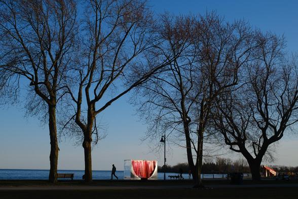 the Slice of Sun winter station, reflecting the setting sun, through two giant trees and a person walking by on the boardwalk