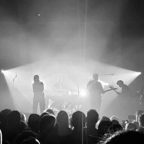 Black and white photo of a live concert scene, showing a band performing on stage with dramatic backlighting and fog effects. Silhouettes of five band members are visible, including a vocalist, guitarist, and keyboard player. The crowd in the foreground is watching attentively. Bright beams of light radiate from behind the band, creating a striking, atmospheric effect.