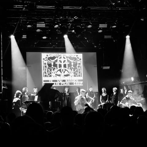 Black and white photo of a live musical performance on a stage, featuring a large ensemble. The group is illuminated by overhead stage lights, with a large screen behind them displaying an ornate, gothic-style image with text that appears to say "IN UW HANDEN [---] MIJN GEEST". The audience is visible in the foreground, watching the performance attentively. The atmosphere suggests a solemn or theatrical theme.