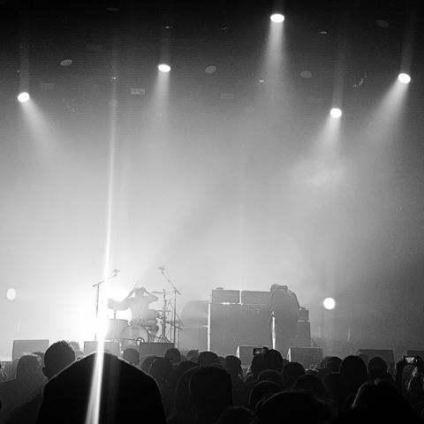 Black and white photo of a moody live concert scene, with a dramatic spotlight beaming from behind the drummer at center stage. The drummer is captured mid-performance, arms raised with drumsticks. Another band member is seen leaning over equipment on the right side of the stage. The atmosphere is heavy with fog and intense lighting, while a large crowd watches in the foreground, some holding up phones to record the moment.
