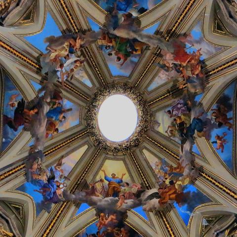 View of a Baroque cupola. In the center is a bright opening through which light enters from the lantern above. The dome is divided into sectors by golden ribs; however, several angels and clouds are painted overlapping these ribs, creating the illusion that they float beneath the dome.