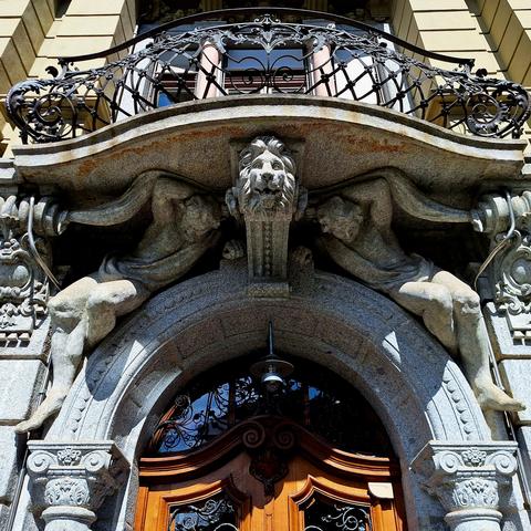 This upward-angled photo shows an ornate entry door. Above the door, the keystone is shaped like a lionhead. Flanking the lion, sculpted human figures support the balcony, which also serves as a roof over the doorway. More information (in German): file:///C:/Users/m.tirassa/Downloads/falkenplatz_22_24_hallerstrass.pdf