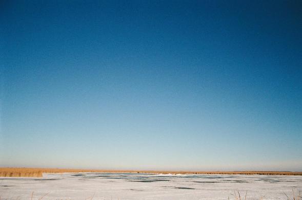 a clear prairie sky, gradient from deep blue to white clouds near the horizon, with a band of golden reeds and white snow on a frozen pond near the bottom of the frame