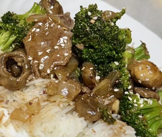 A close up of beef, broccoli, mushrooms, and onions on rice sprinkled with sesame seeds and dried chili peppers, on a white plate. 
