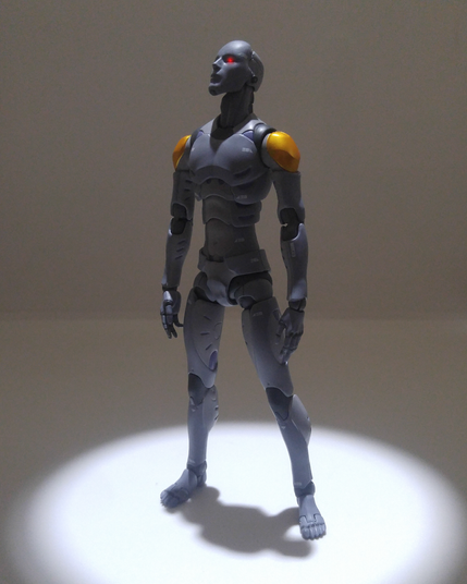 A 1/12 scale TOA Heavy Industries Synthetic Human E.S.G.S model 3 standing under a spotlight in a dark room with white walls. Its eyes are glowing red.