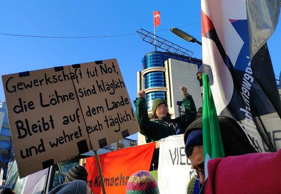 Bild einer Demonstration. Darauf ein Lautsprecherwagen und Fahnen der Gewerkschaftsföderation FAU. Im Vordergrund ein Schild mit der Aufschrift: "Gewerkschaft tut Not, die Löhne sind kläglich. Bleibt auf dem Lande und wehret euch täglich!"