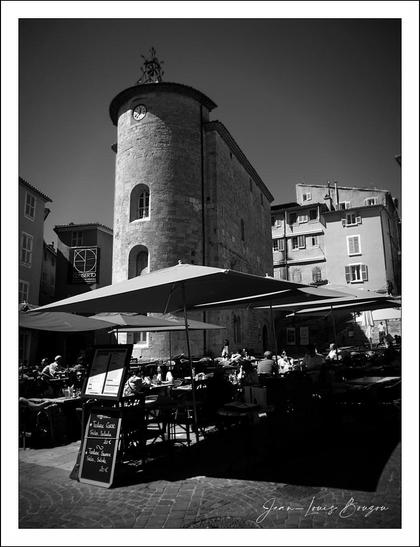 The image presents a charming urban scene captured in black and white, emphasizing texture and contrast. In the foreground, a lively outdoor café is bustling with patrons gathered under large umbrellas, enjoying the day. Tables are neatly arranged, some adorned with menus written in chalk, hinting at a vibrant culinary offering.
The backdrop features a tall, stone tower, which has a cylindrical shape and is characterized by its rustic, weathered façade. The tower stands tall against a clear sky, accentuating its historical significance and architectural solidity. Windows adorned with simple frames punctuate the stonework, and the structure's upper edge is crowned with a small decorative element, adding a quaint charm.
Surrounding buildings are visible, adding depth to the composition, with their facades slightly blurred in the background while the café scene remains crisp and inviting. The interplay of shadows and sunlight enhances the inviting atmosphere of this lively square, evoking a sense of community and the joys of outdoor dining. The absence of color allows for a focus on form and light, enriching the emotional resonance of the moment captured.
Overall, the image evokes a serene yet lively ambiance, celebrating both the architecture and social interaction typical of bustling squares in historic towns.
