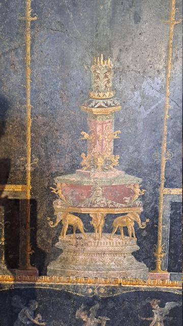 Photo of a Pompeii Roman wall painting detail which is rather hard to decipher but it looks rather like a wedding cake... a pillar or decorative monument with several sections getting wider towards the bottom with the second to last section held up by three golden elephants holding something in their trunks, all standing on a marble pedestal... it looks like it was influenced by eastern or indian art but perhaps elephants are more common than I thought in Roman art