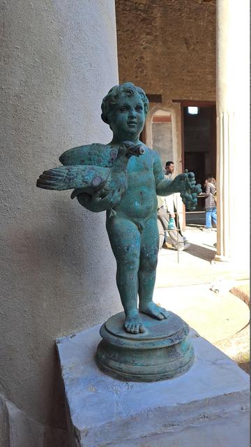 Photo of a Pompeii Roman villa metal statue of a young boy or cherub, naked, holding a bird like a duck in one arm and a bunch of cherries in the other hand