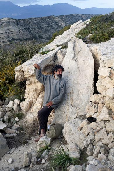 Un homme à la barbe poivre et sel, pieds nus, se se tient appuyé sur une grosse pierre: le tête tournée vers la droite de la photo: il regarde en direction l’entrée de la grotte qu’il habite. Au loin, de hautes montagnes. 