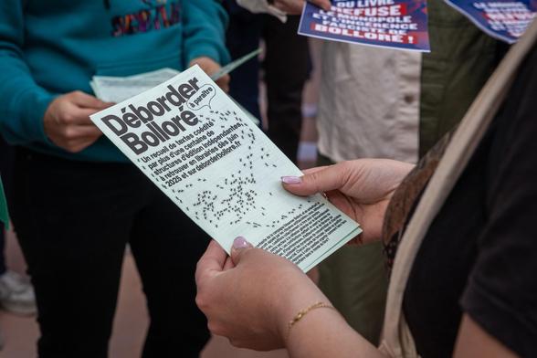Photographie du communiqué de presse Déborder Bolloré tenu par deux mains, tract en arrière plan indiquant "Le monde du livre refuse le monopole et l'ingérence fasciste de Bolloré"
