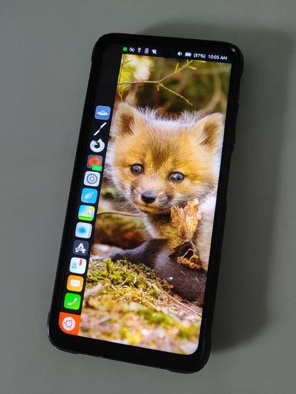 Photo of a Poco X3 NFC smartphone running Ubuntu Touch showing app dock on left edge with a wallpaper of a fox cub on screen.