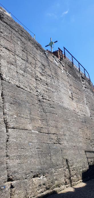 Das Bild zeigt eine massive, steinerne Mauer, die sich in einem hohen Winkel nach oben erstreckt. Die Mauer besteht aus großen, unregelmäßig geformten Steinblöcken, die in verschiedenen Grautönen gehalten sind. An der Oberseite der Mauer befindet sich ein Metallgeländer, das wahrscheinlich als Sicherheitsmaßnahme dient. Auf dem Geländer steht ein Kreuz, das mit bunten Fäden geschmückt ist. Der Himmel ist klar und blau, was auf einen sonnigen Tag hinweist. Die Mauer weist einige Risse und Verwitterungserscheinungen auf, was auf ihre Alter und Belastung durch die Elemente hinweist.