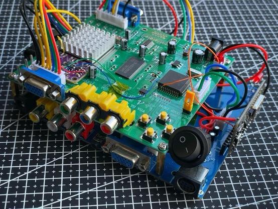 A close-up view of a green printed circuit board (PCB) with various electronic components, including multiple connectors, a heat sink, and colorful wires. The board features input/output ports, buttons, and capacitors, all positioned on a grid-patterned