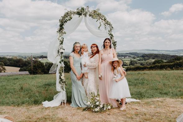 Posed group wedding photo of the bride, bridesmaids and flower girls standing under a floral arch during an outdoor handfasting ceremony. The scene captures a soft, feminine atmosphere with pastel dresses, natural countryside light and wildflowers in bloom. This image reflects the elegant, unposed beauty of documentary wedding photography – where even formal moments feel relaxed and authentic. A visual story from a small, heartfelt wedding in the countryside, focusing on sisterhood, connection and candid storytelling through photography.