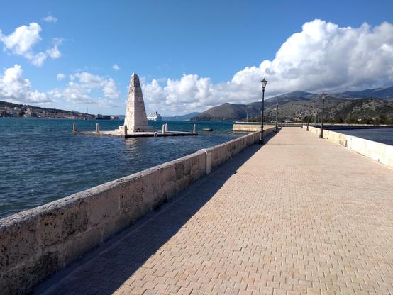 Picture of bridge, now paved and pedestrianised, stretching across the lagoon. At the bend in the bridge is an obelisk and the bay beyond surrounded by hills. In the distance is a departing cruise ship. 