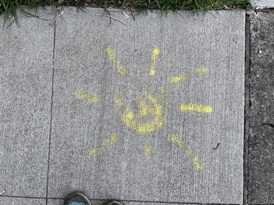 Photo of a yellow sun in paint or maybe chalk on the sidewalk. The toes of my beat-up sneakers are barely visible at the bottom of the image. 
