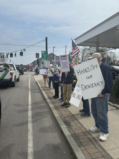 A group of protesters stands along the roadside, holding signs that express various political messages, including "Dump Trump" and "Hands Off Our Democracy." Some individuals are also waving American flags. The setting features traffic signals and a gas station in the background.