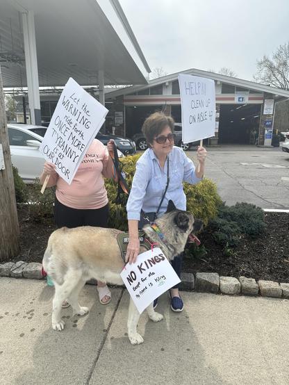 Two women are standing outside holding signs with messages. One sign reads "Let the WARNING ride forth ONCE MORE Tyranny is at OUR DOOR," and the other says "HELP!!! CLEAN UP on AISLE 47." An Akita dog is wearing a sign saying “No Kings, except for me. My name is King”.