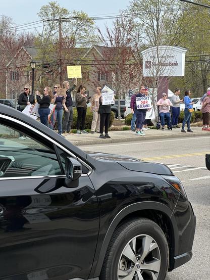 A group of demonstrators stands on the sidewalk holding signs advocating for veterans' support. They appear engaged and passionate about their cause. 