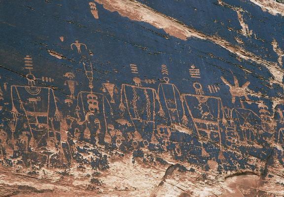 Six pecked human-like forms with large headdresses on a dark cliff wall, as well as other smaller forms.