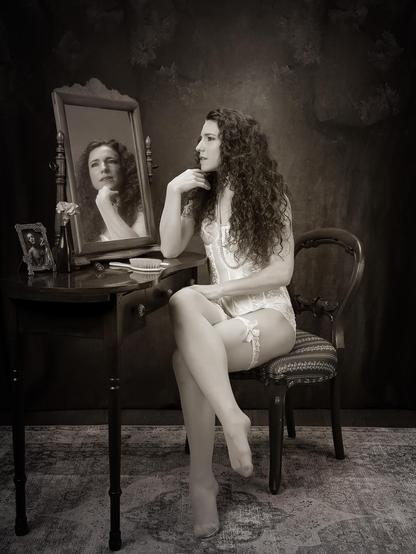 Toned black and white photo of model Keira Grant poses in white lingerie and stockings in front of a dressing table.  She is gazing into a mirror on the table, and her reflection can be seen in the mirror.