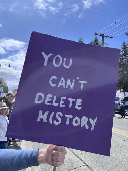 Phot of a sign at a protest: purple poster board with silver lettering: YOU CAN’T DELETE HISTORY
