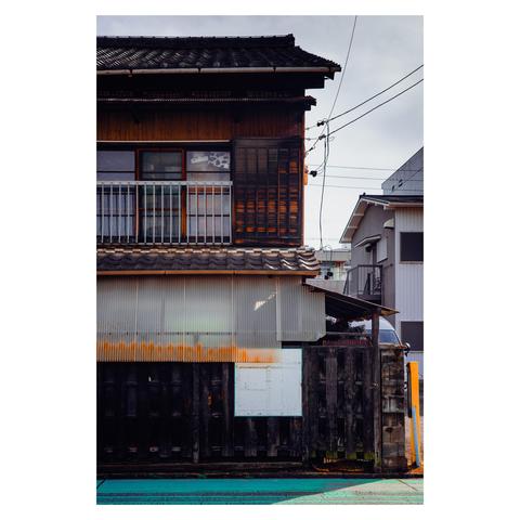 A traditional Japanese house, its darkened timber siding and tiled roof softened by the season’s gentle sunlight. Corrugated plastic sheets hang slightly uneven, catching the warm glow. The faint green of a painted curb below hints at new growth, while utility wires stretch above like quiet lines in a springtime sketch.
