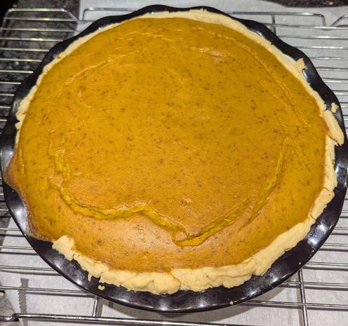 A large pumpkin pie l, just out of the oven sitting on a cooling rake. There are cracks showing on top, and some filling overflowed the crust in one part 