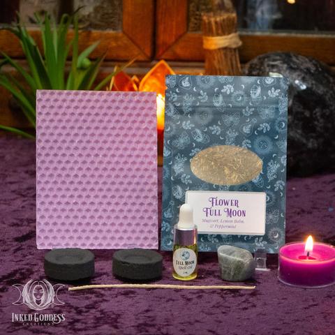 The contents of our exclusive "Flower Full Moon Ritual Kit for the May 12, 2025, Full Moon in Scorpio" product are arranged on a velvet-covered surface. They include a sheet of purple beeswax and a wick; a pouch of Flower Full Moon herbs, including mugwort, lemon balm, and peppermint; a dropper vial of our exclusive Full Moon Spell Oil; a tumbled labradorite gemstone; and two charcoal discs. Presented by Inked Goddess Creations.