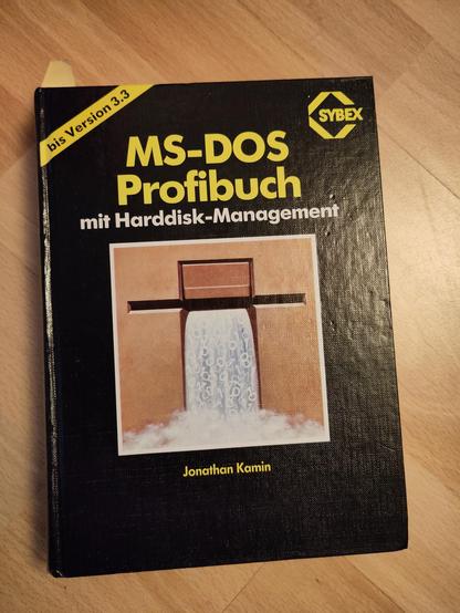Das Bild zeigt ein Buch mit dem Titel "MS-DOS Profibuch mit Harddisk-Management" von Jonathan Kamin. Das Buch hat ein schwarzes Cover mit gelben und weißen Buchstaben. Im oberen linken Bereich des Covers befindet sich ein gelbes Streifen mit der Aufschrift "bis Version 3.3". Der Titel des Buches ist in großen, gelben Buchstaben gedruckt, und darunter steht in kleineren weißen Buchstaben "mit Harddisk-Management". Der Name des Autors, Jonathan Kamin, ist in gelben Buchstaben am unteren Rand des Covers zu sehen. Das Cover zeigt ein Bild eines Wasserfalls, das von einem Brett durchlaufen wird, und symbolisiert möglicherweise Datenfluss oder Informationsspeicherung. Das Buch ist von der Verlagsserie Sybex, wie das gelbe Sybex-Logo im oberen rechten Bereich des Covers zeigt. Das Buch liegt auf einem Holztisch.