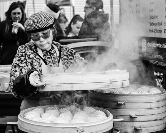 Black and white image of a woman wearing sunglasses while tending to hum bao in bamboo steamers