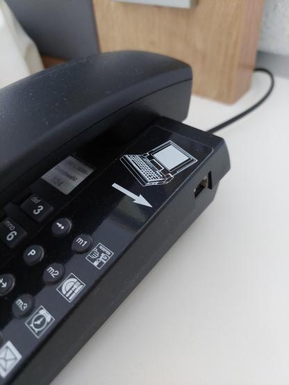 Man sieht ein schräg von der Seite fotografiertes Tasten Festnetztelefon. Neben den Nummerntasten sieht man Tasten, die für den Hotelbetrieb bestimmt sind (u. A. Rezeption, zimmerservice, Weckdienst,...). Oben rechts sieht man ein Pictogramm eines Laptops (ca. Mitte der 90er) mit einem Pfeil zur Seite des Telefons. An der Seite des Telefons befindet sich ein Anschluß für ein Modem.