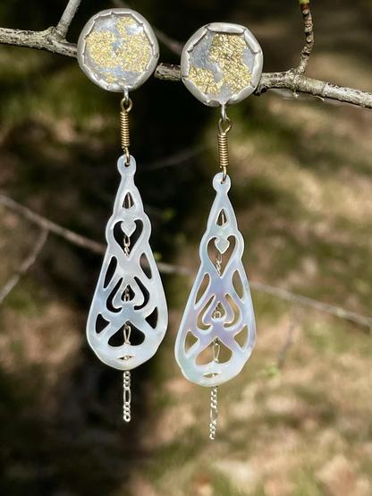 A pair of elegant earrings showcasing circular tops with a glittery finish, connected to intricate, elongated teardrop shapes made of a translucent material. The earrings hang from a branch, set against a blurred natural background.