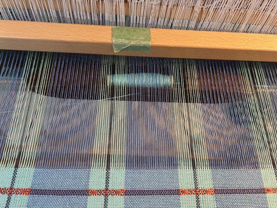 A wooden boat shuttle with light blue cotton midway through a pass while weaving, partially hidden by warp threads