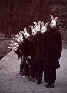 zwartwitfoto van een rij mensen die schuin achter elkaar staan, steeds iets verder opzij buigend om zichtbaar te zijn voor de fotograaf. Ze dragen allen een masker van een haas.