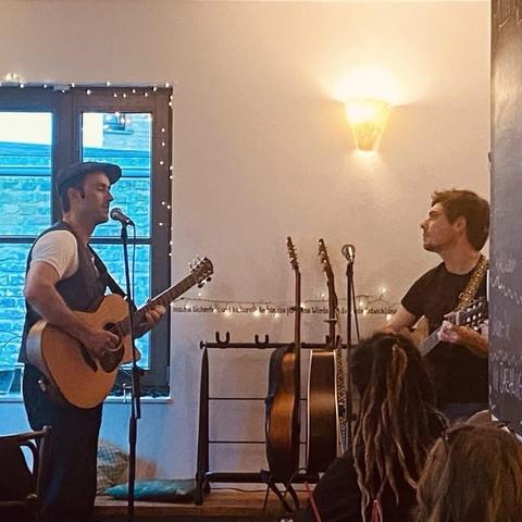 Zwei Musiker treten auf der Bühne in einem gemütlichen Veranstaltungsort auf. Der eine spielt eine Akustikgitarre und singt in ein Mikrofon, während der andere ihn mit einer anderen Gitarre begleitet. Mehrere Gitarren sind im Hintergrund zu sehen, zusammen mit warmer Beleuchtung und einer entspannten Atmosphäre.