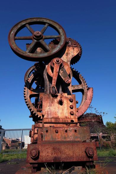 Stählernde Metallkonstruktion mit Hebeln, Zahnrädern und Riemenscheiben für den Antrieb. Das rostrote Maschinenteil hebt sich vor einem tiefblauen Himmel (nachbearbeitet) ab.

Ursprünglich diente es in der Stahlverarbeitung. Heute erinnert es an eine Stahlskultpur.