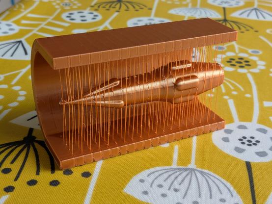 A shiny 3d printed model, made from bronze coloured "silk" filament. It's a model of the ship "Limiting Factor" from the Iain M Banks book "The Player of Games". The ship is shaped like a kind of spindle, tapering to a point at the nose with a bulge at the waist and a bluff truncated cone at the rear. Leading up to the nose there are three long bulges, with five shorter ones around the waist. The model is printed inside a C-shaped holder, suspended by thin filament strands so it looks a little like it's flying