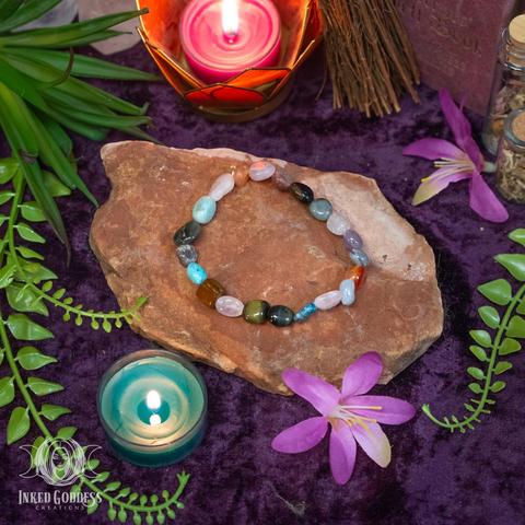 An example of the exclusive Mixed Tumbled Gemstone Bracelet sits atop a flat red rock. The bracelet is elastic and has no clasp. It's made of small irregularly-shaped polished gemstone beads, including rose quartz, agate, aventurine, and more. This is an exclusive product from Inked Goddess Creations.