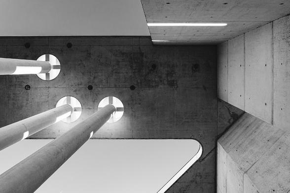 Black and white upward view of a modern concrete structure featuring large circular openings with cylindrical columns passing through them. The composition showcases geometric precision and dramatic lighting, blending architectural minimalism with abstract design.

