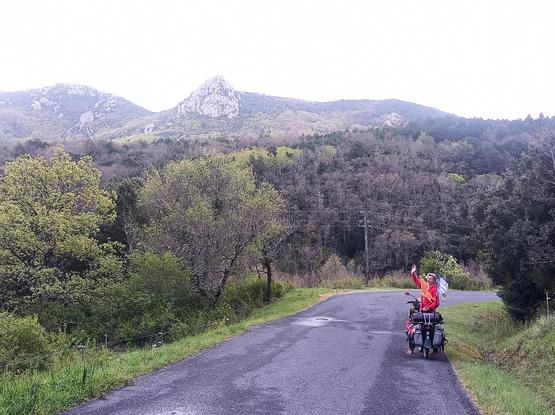 A cyclist waving going down a hill, green shrubs and trees and mountains in the background
