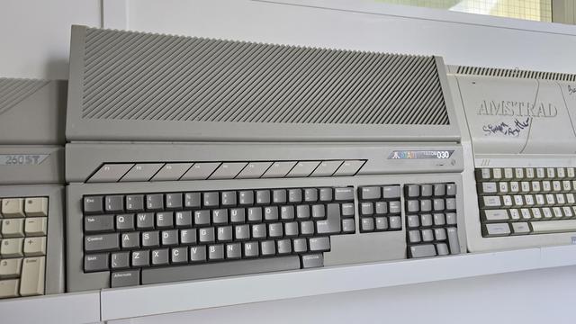 Photograph of a grey Atari Falcon030 computer on display, flanked by an Atari 260ST on the left and an Amstrad CPC model on the right. The Falcon030 features a distinctive diagonal vent design, dark grey function keys (F1–F10), and a full-size keyboard with a numeric keypad. The Atari and Falcon030 logos are visible above the keyboard. The Amstrad machine to the right bears a visible signature, adding a personal collector's touch. All three computers are arranged on a white shelf.