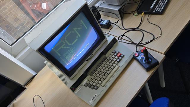 Tatung Einstein computer with built-in monitor displaying the “X BASIC MENU”. A black joystick with red buttons labelled “Super” is connected. A ZX Spectrum is also visible in the background, with cables running to a small screen. The setup is arranged on a wooden desk by a window.