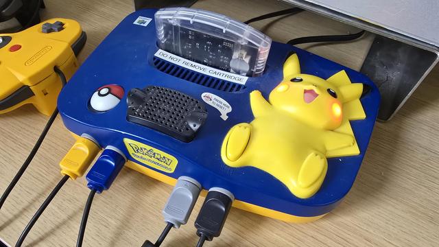 Pokémon-themed Nintendo 64 console featuring a large yellow Pikachu figure with glowing red cheeks. The reset button is integrated into Pikachu's foot, and a raised Poké Ball button is on the left. A translucent cartridge labelled “DO NOT REMOVE CARTRIDGE” is inserted. Four controller ports are connected at the front, and a yellow Pokémon-branded controller is visible to the left. The console rests on a wooden surface.