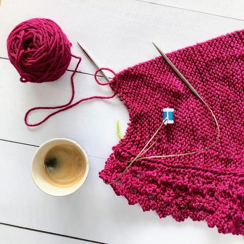 Flat lay view of a ball of dark pink yarn, and a knitting project featuring garter stitch and lace on a circular needle. It's shown on a white plank background, together with a cup of coffee.