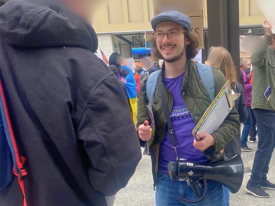 A FairVote Illinois volunteer with a clipboard and a bullhorn draped around his waist smiling and talking with a rally attendee. 