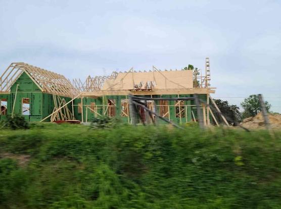 Unfinished house under construction, wooden frame, green sheathing, surrounded by grassy field.